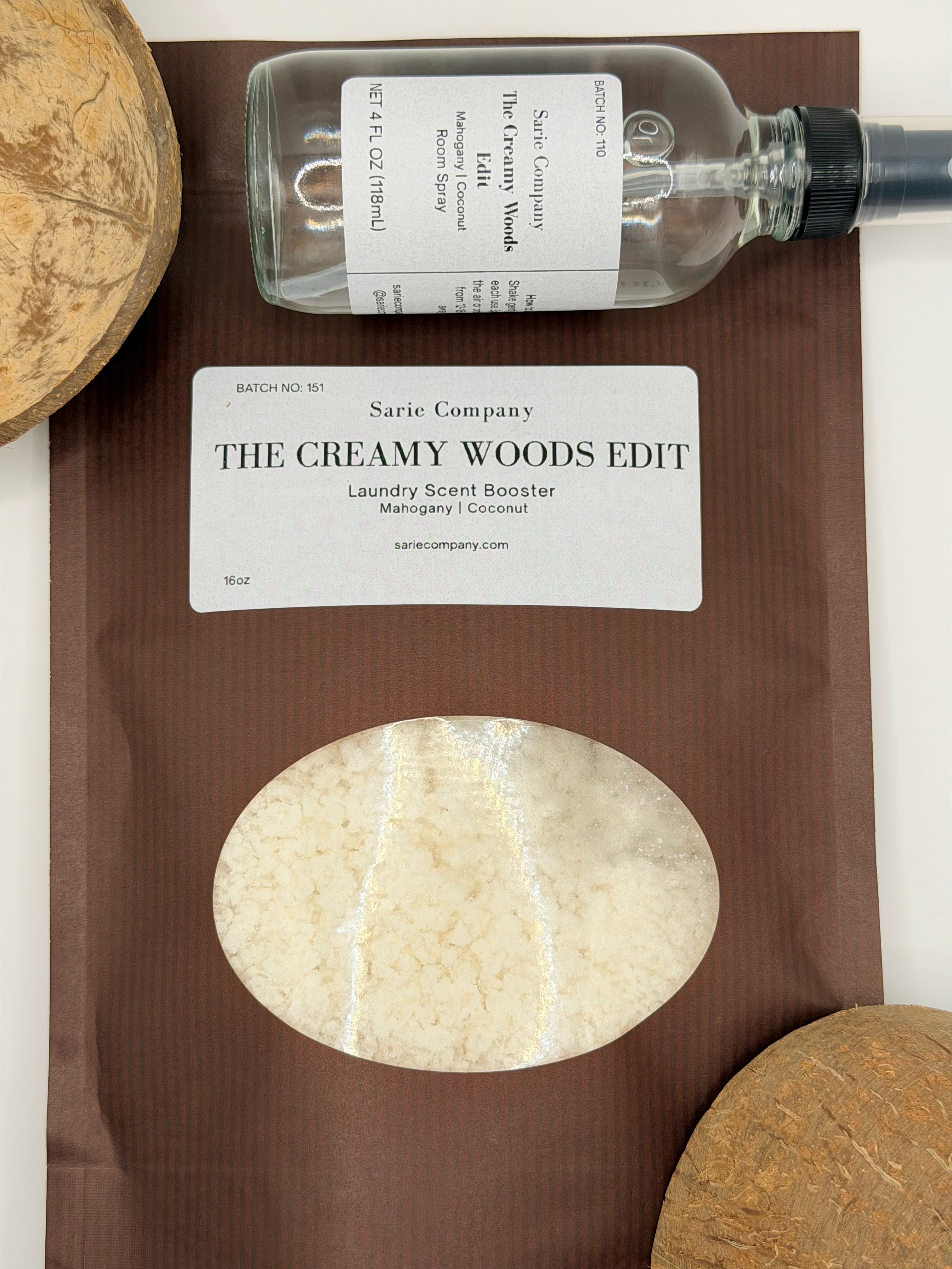 A room spray and laundry scent booster from Sarie Company's Creamy Woods Edit displayed with coconuts on a white surface.