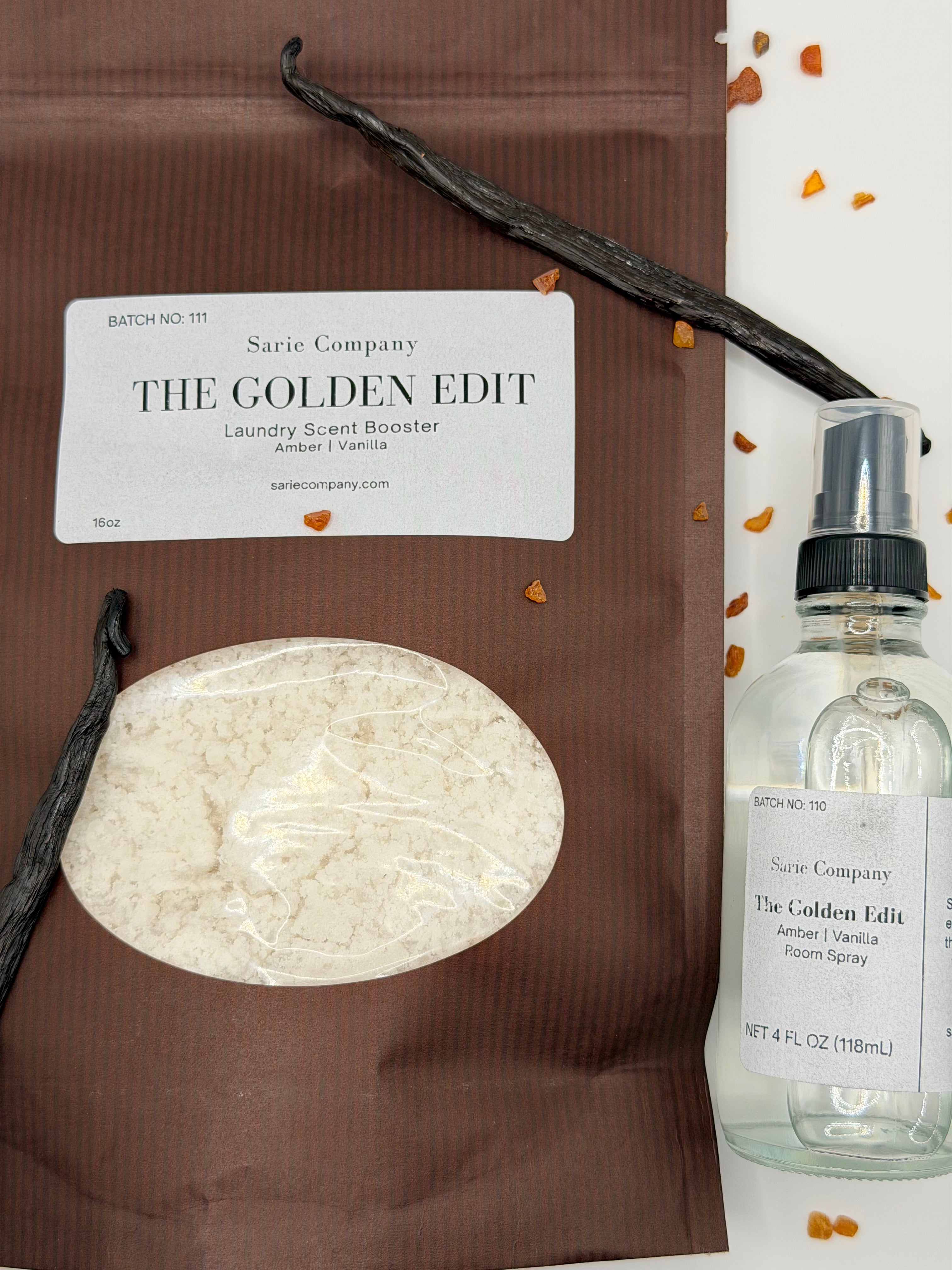 A room spray and laundry scent booster from Sarie Company's Golden Edit displayed with vanilla beans, and amber crystals on a white surface.