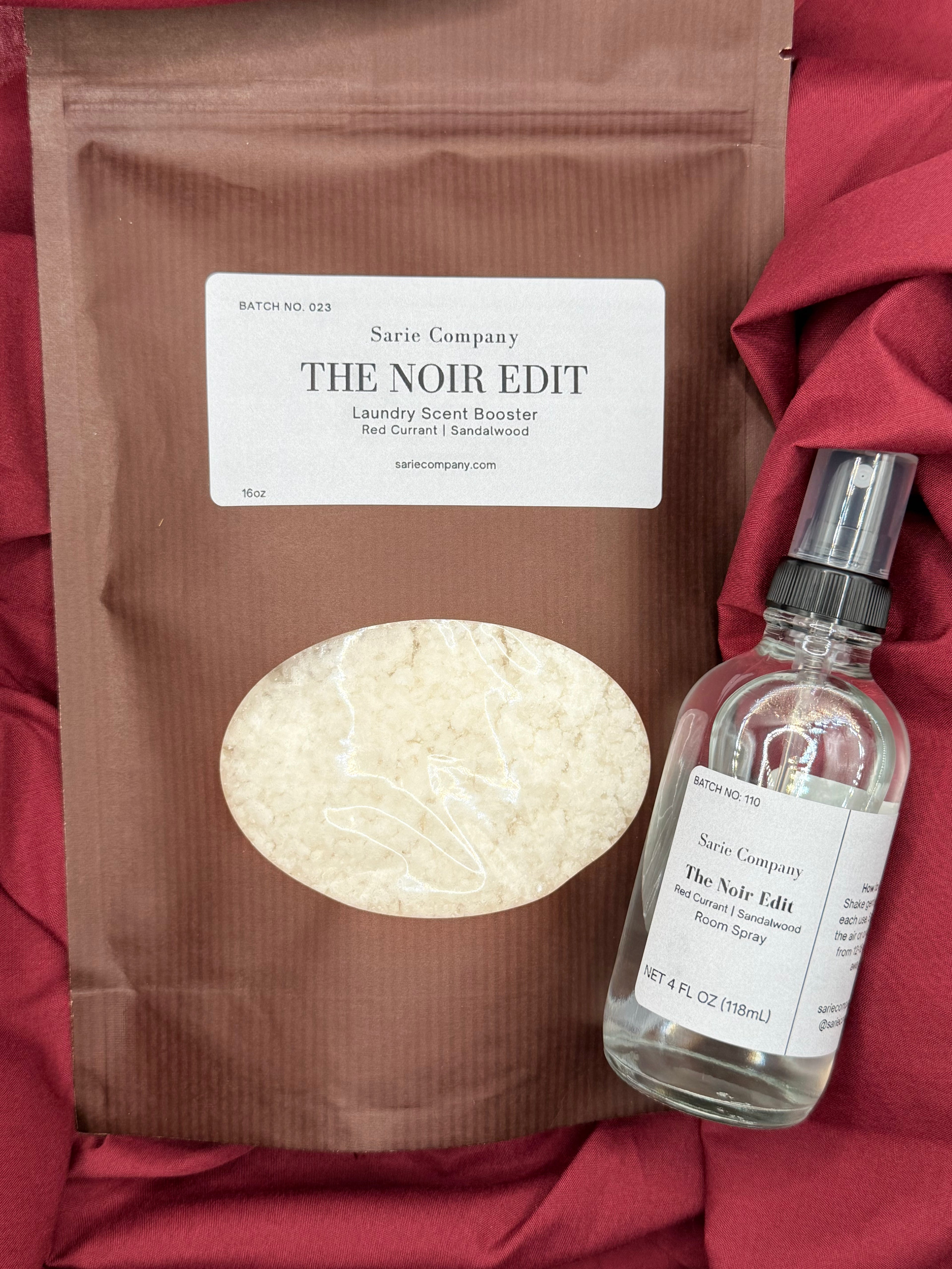 A striped brown resealable pouch and a clear room spray bottle from Sarie Company's The Noir Edit collection displayed on a red fabric background. The pouch contains a luxury scent booster, and the spray bottle is labeled as a room spray. Both items feature matching white labels and a warm, elegant aesthetic.