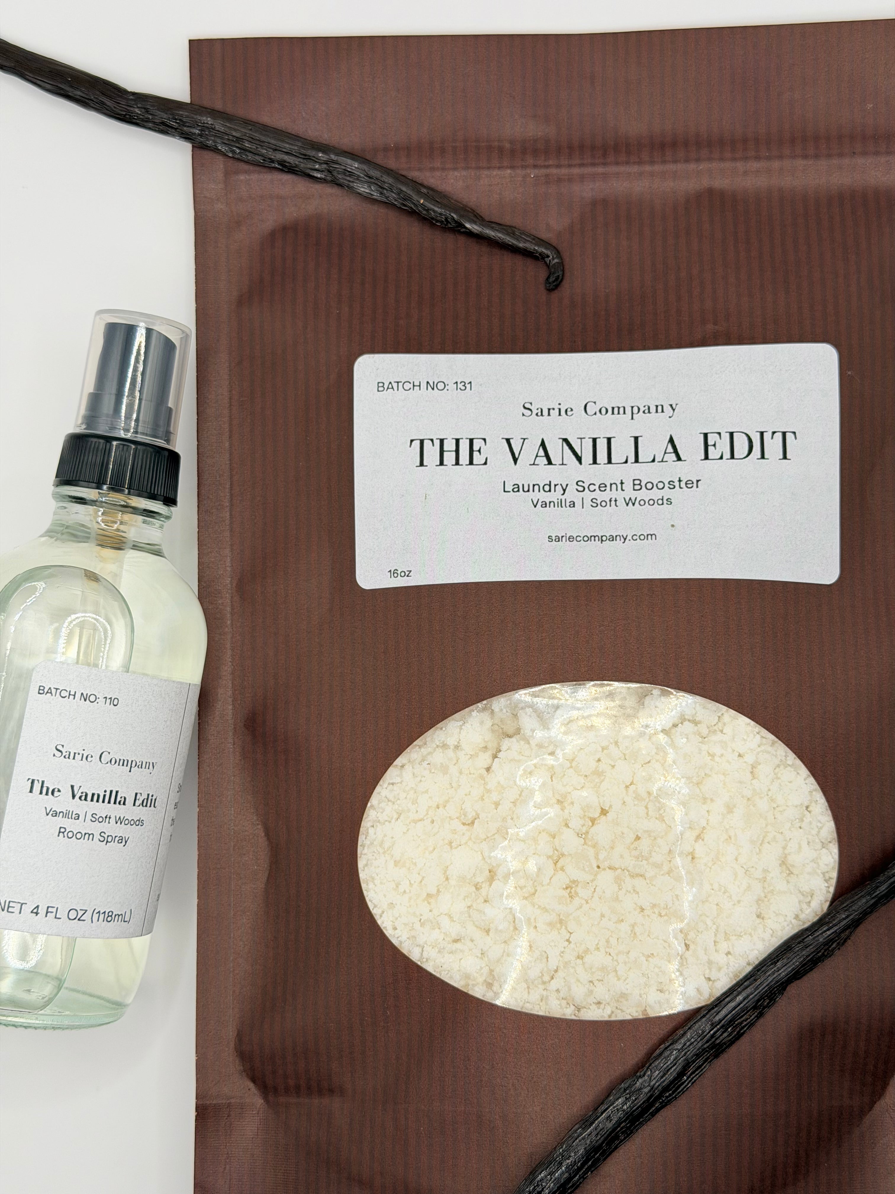 A room spray and laundry scent booster from Sarie Company's Vanilla Edit displayed with vanilla beans on a white surface.